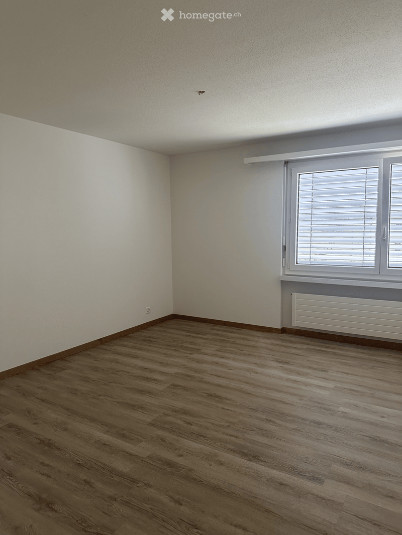 Empty room with wooden flooring, white walls, and a large window with blinds. A radiator is visible below the window.