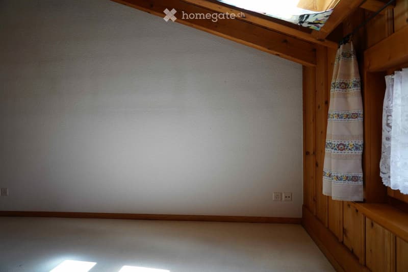 Empty room with wooden beams, white walls, and a skylight allowing natural light to enter. The floor is carpeted, and there are wooden shelves and a curtain visible.