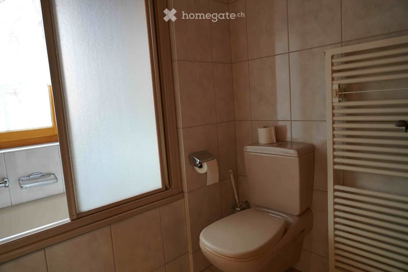 A small bathroom with beige tiles, a toilet, toilet paper holder, a radiator, and a frosted window.