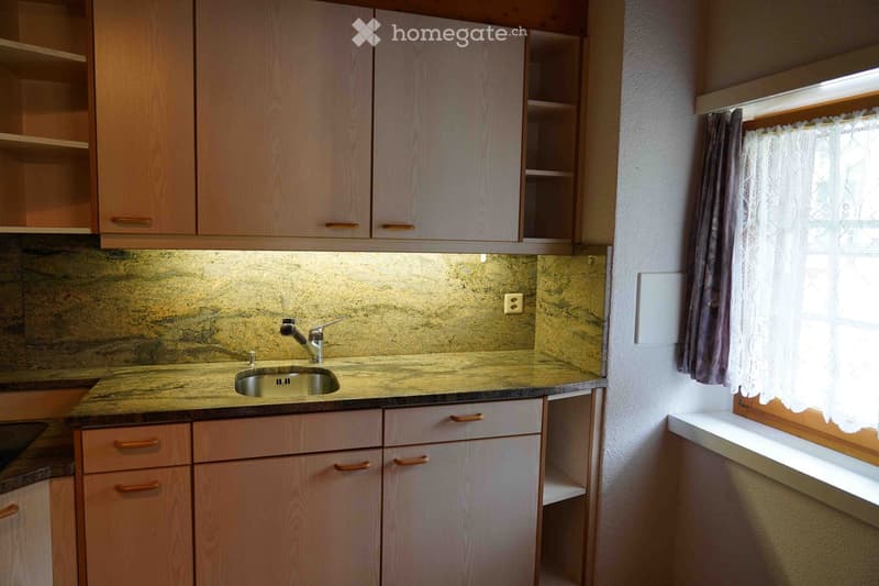 A small kitchen with light wooden cabinets, a marble countertop, and a sink with a faucet. The backsplash is made of a textured beige material, and there is a window with curtains on the right side.