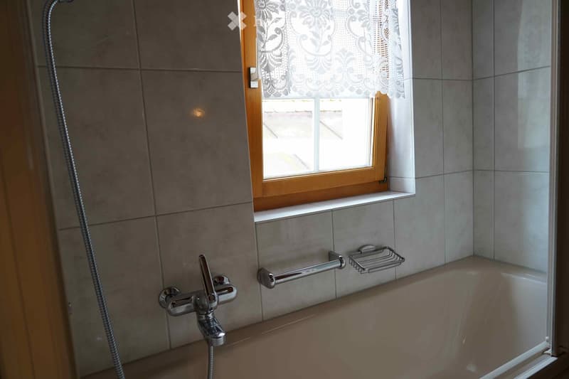Bathroom with tiled walls, a bathtub, a shower hose, a faucet, a handrail, and a towel rack. A window with a lace curtain is above the bathtub.