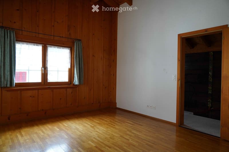 Empty room with wooden walls and flooring, large window with curtains, and an open doorway leading to another room.