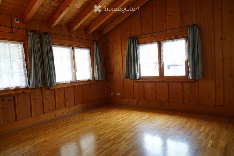 Empty wooden room with exposed beams, large windows with curtains, and wooden flooring.