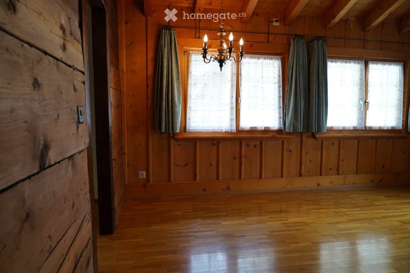 Empty room with wooden walls and floor, large windows with curtains, and a chandelier hanging from the ceiling.