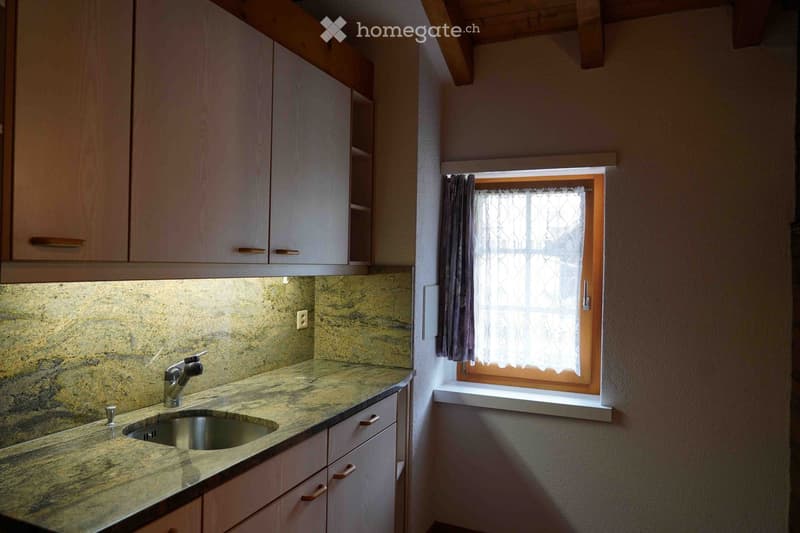 A small kitchen with a granite countertop, a sink, and wooden cabinets. A window with a curtain lets in natural light.