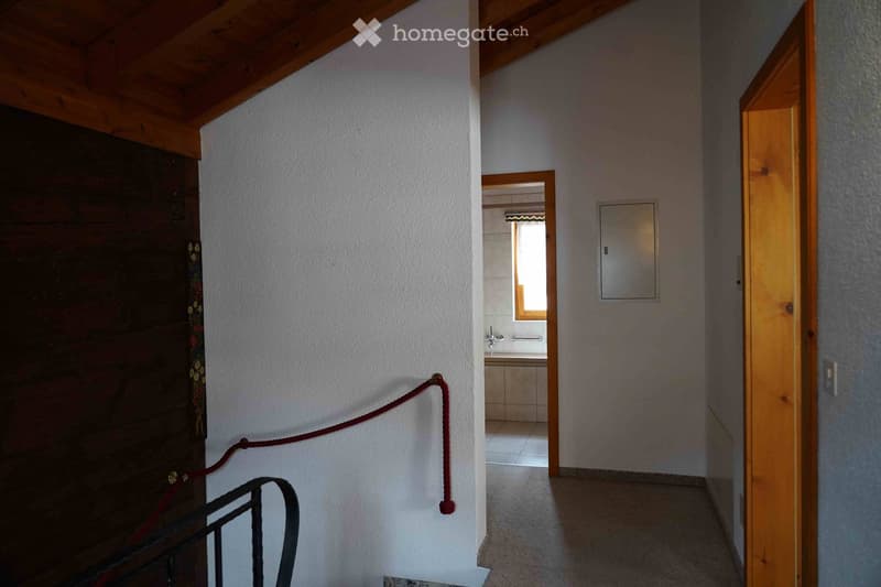 A narrow hallway with wooden doors, a visible bathroom with a window, and a metal railing on the left side.