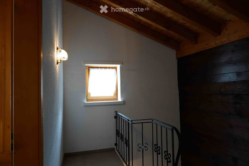 A narrow hallway with wooden beams on the ceiling, a small window with white frame, and a metal railing leading upwards.