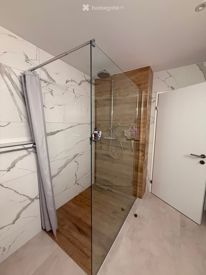 Modern bathroom with a glass-enclosed shower, marble-patterned tiles, wooden floor, and a shower curtain.