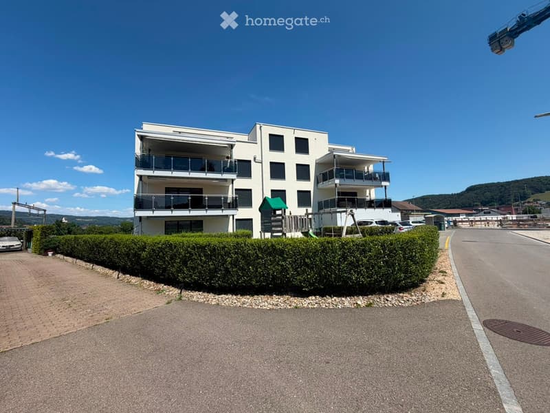 Modern two-story residential building with balconies, surrounded by a neatly trimmed hedge and a paved driveway.