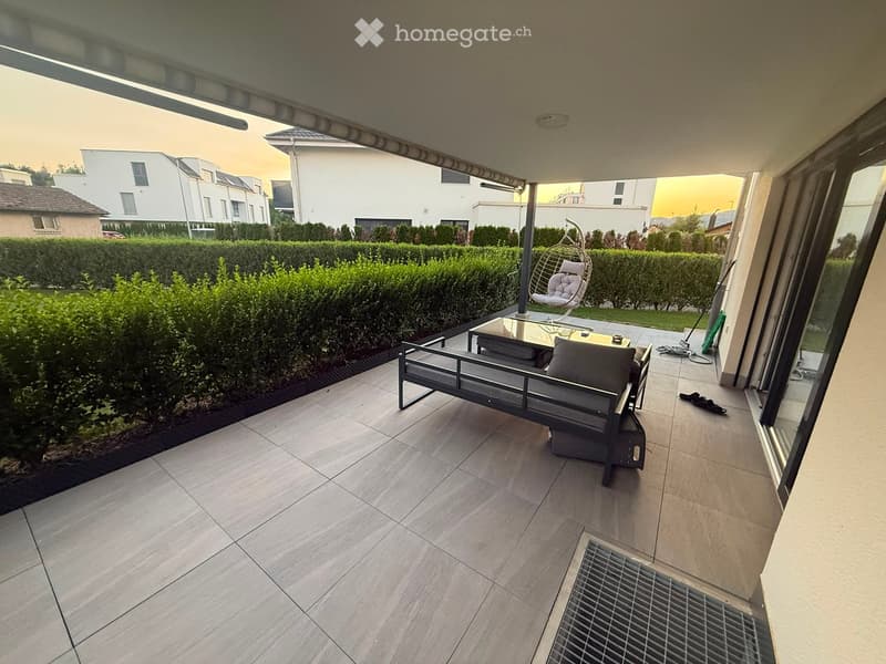 Balcony with tiled floor, sofa, table, and chair, surrounded by neatly trimmed hedges and neighboring houses.