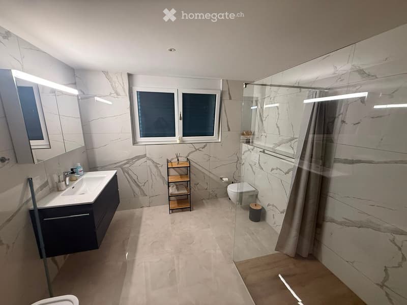 Modern bathroom with marble tiles, a large window, a sink with a dark cabinet, a toilet, a shower with a glass enclosure, and a small wooden towel rack with towels.