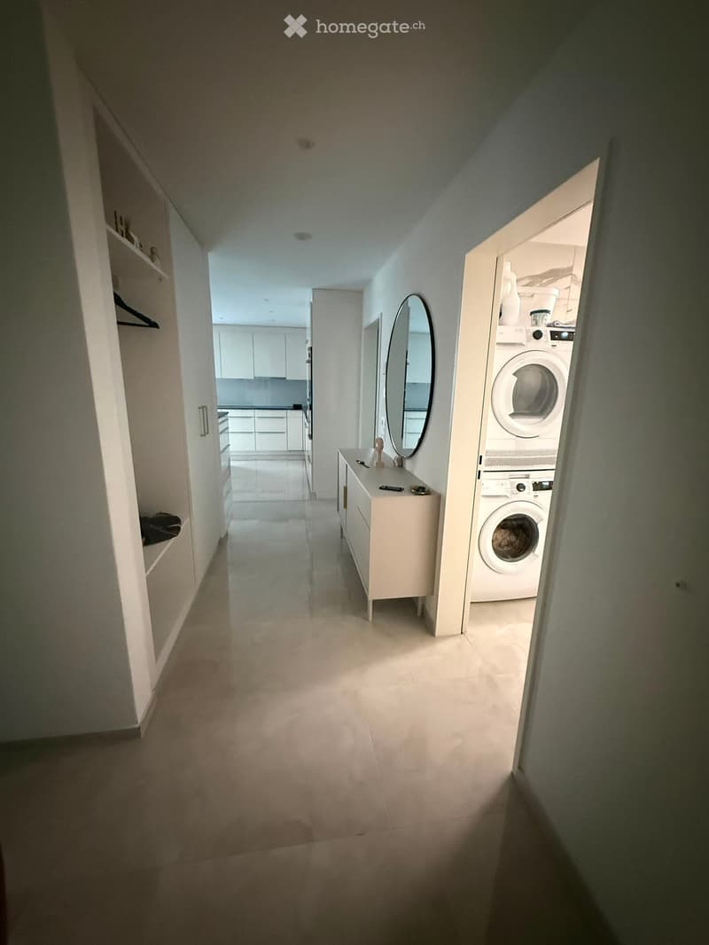 A narrow hallway with white walls, a small shelf on the left, and a small table with a mirror and a few items on the right. The hallway leads to a kitchen area with white cabinets and a washing machine in the background.