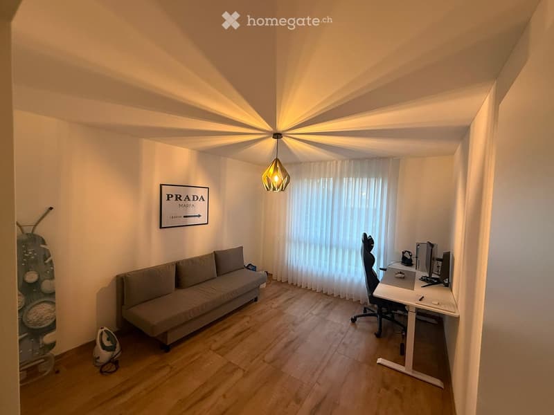 A modern, well-lit room with a sofa, desk, chair, laptop, and a hanging lamp. The room features wooden flooring, white walls, and a large window with curtains.