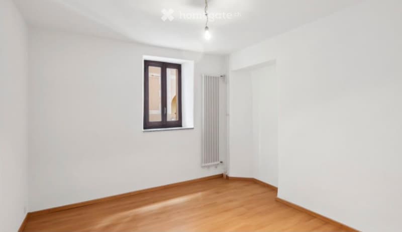 Empty room with white walls, wooden flooring, a single window with dark frame, radiator, and a ceiling light fixture.