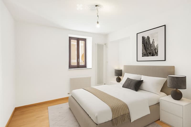 Modern bedroom with a large bed, nightstands, lamps, and a framed picture on the wall. The room features wooden flooring and large windows.