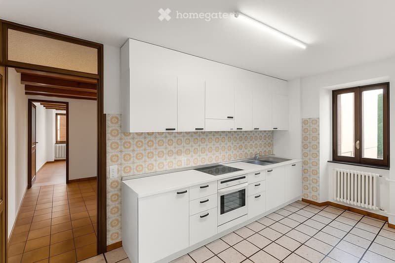 Modern kitchen with white cabinets, a sink, an oven, and a tiled backsplash. The space features a window and a hallway leading to another room.