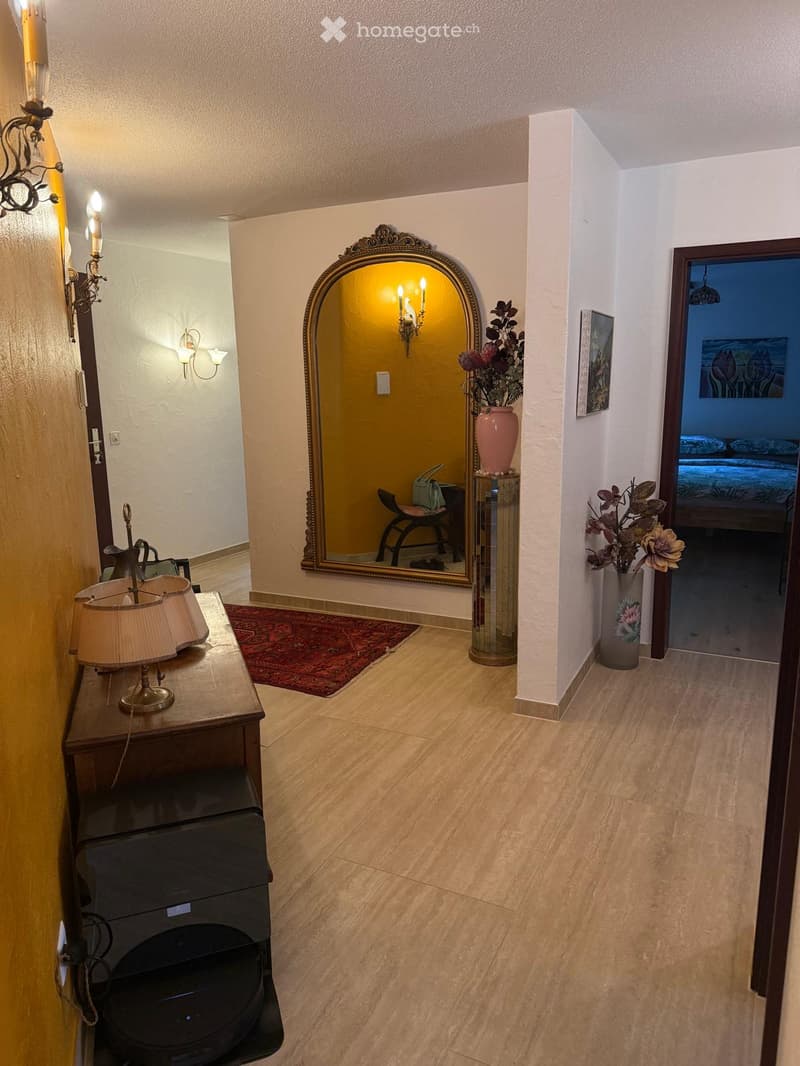 Elegant hallway with wooden flooring, a large ornate mirror, a table with a lamp, and vases with flowers. A doorway leads to a bedroom.