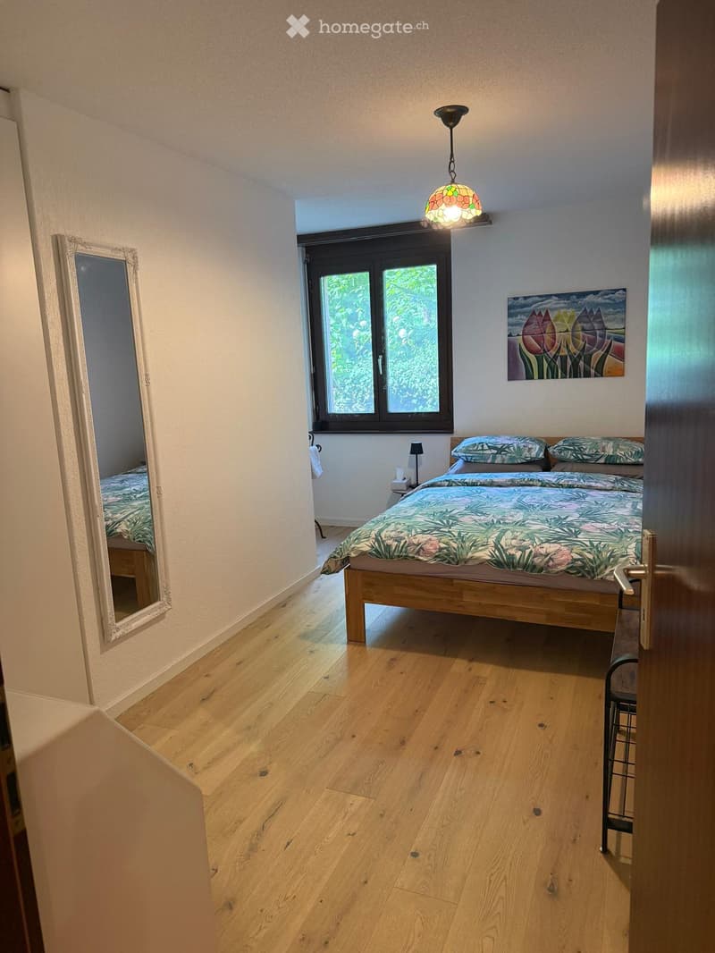 Cozy bedroom with wooden flooring, a double bed with a colorful duvet, a mirror, a small table lamp, and a painting on the wall.
