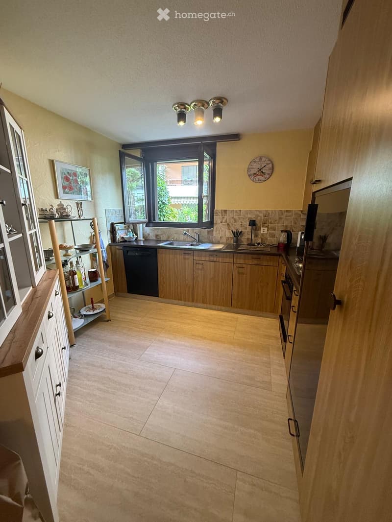 Bright kitchen with wooden cabinets, tiled floor, and a large window. Includes a sink, dishwasher, oven, and various kitchen items on shelves and counters.