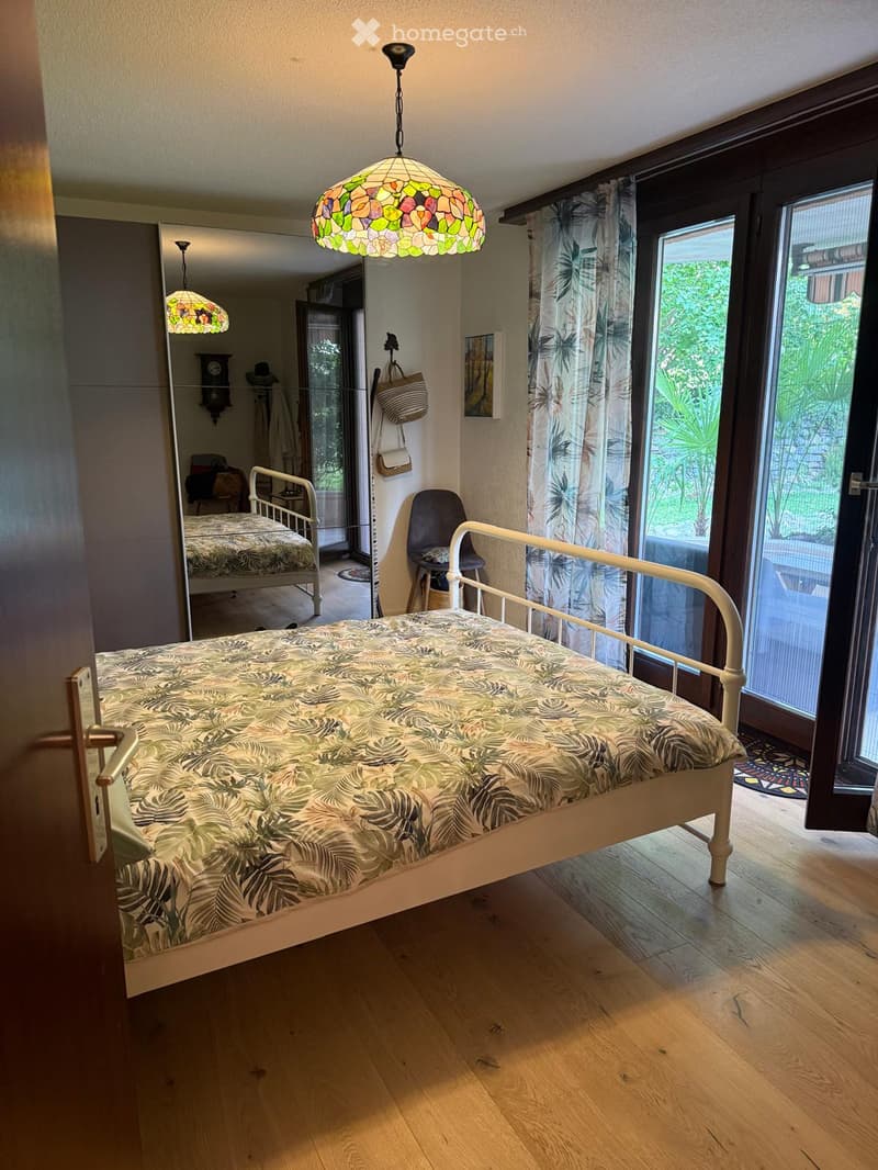 Bright bedroom with wooden flooring, a single bed with a leafy-patterned duvet, a hanging stained-glass lamp, a chair, a mirror, and a view of greenery through large windows.