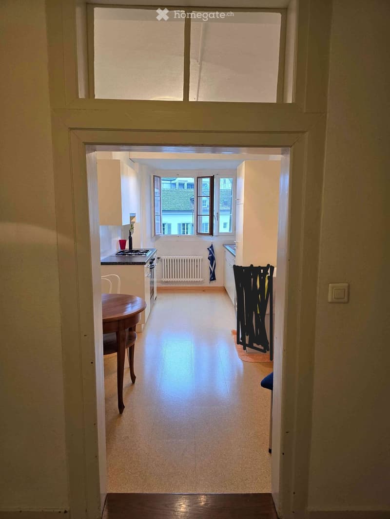 A narrow hallway with light-colored walls and a polished floor, leading to a small kitchen area with a stove, sink, and cabinets.