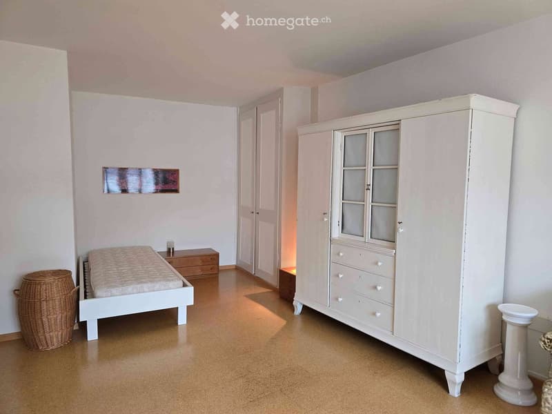A well-lit bedroom with a white bed, a large wardrobe with glass doors, a nightstand, and a wicker basket. The room features light-colored walls and a wooden floor.