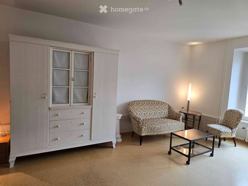 A well-lit living room with a patterned sofa, matching armchair, glass coffee table, and a large white wardrobe with glass-paneled doors.