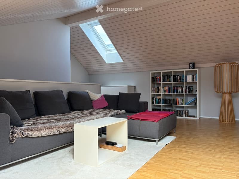 Modern living room with grey sectional sofa, fur throw, coffee table, bookshelf, and floor lamp. Wooden ceiling with skylight.