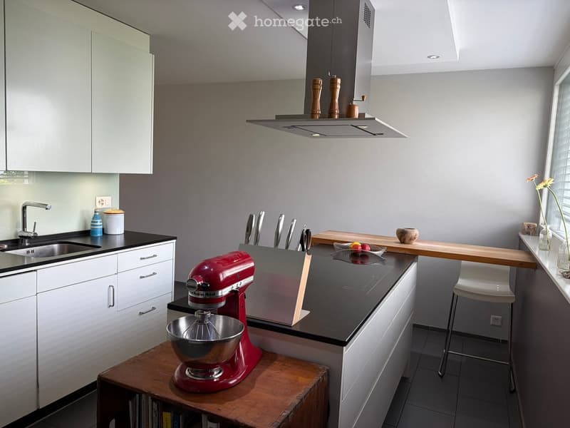 Modern kitchen with white cabinets, black countertops, a red stand mixer, and a wooden island with a bowl of fruit. Features a stainless steel sink, faucet, and a pepper grinder on the backsplash.
