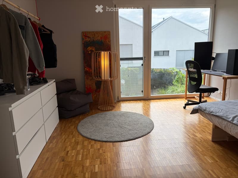 Modern bedroom with wooden flooring, a round grey rug, a bed with grey bedding, a beige sofa, a desk with a chair, a floor lamp, and a drawer unit with hanging clothes.