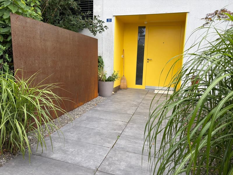 Modern outdoor area with a concrete pathway leading to a bright yellow front door, surrounded by greenery and a metal fence.