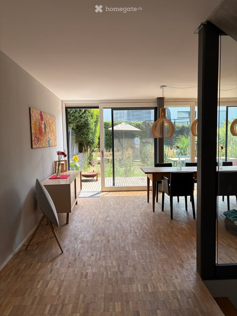 Bright dining area with wooden flooring, large glass doors opening to a garden, a dining table with chairs, a sideboard with a vase, and a modern lamp hanging from the ceiling.