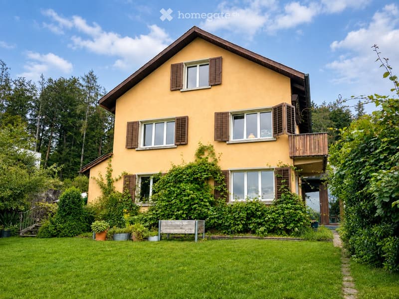 Two-story yellow house with brown shutters, surrounded by greenery and a well-maintained lawn. Features a balcony on the upper level and a small wooden bench in the front yard.