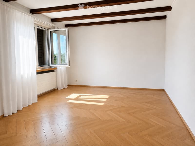 Empty room with light wooden herringbone flooring, white walls, exposed wooden ceiling beams, and large windows with white curtains.
