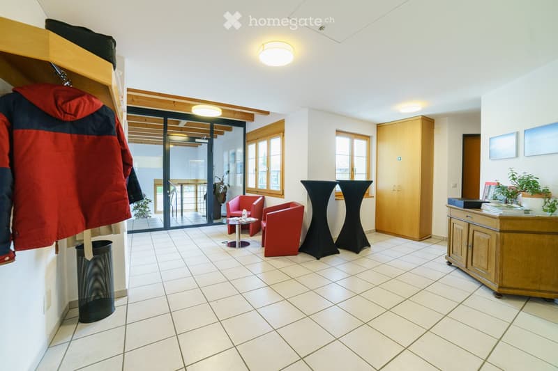 Bright, modern room with white tiled flooring, large windows, and a sliding glass door leading to a balcony. Features include red chairs, a small table, tall black stools, a wooden cabinet, and a coat hanging on the wall.
