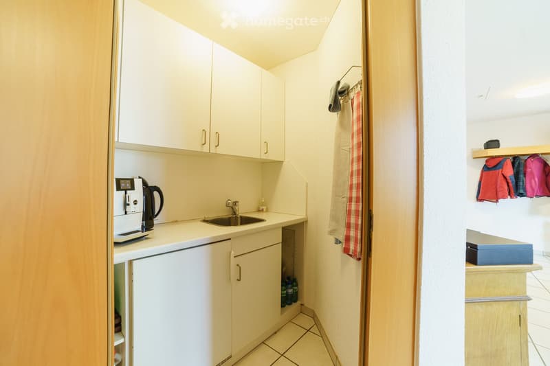 Compact kitchen with white cabinets, a coffee maker, a sink, and a small storage area with hanging clothes and a shelf.