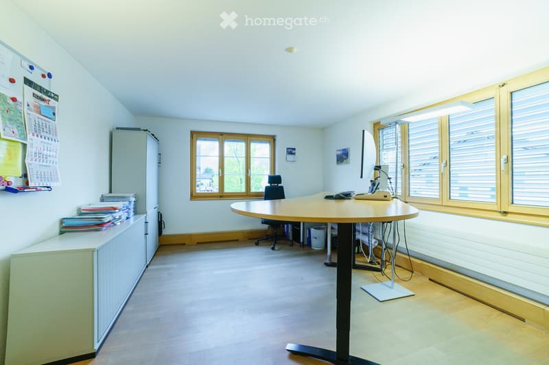 Bright office space with a large wooden table, office chair, filing cabinet, calendar, and computer screen. Natural light from large windows with wooden frames.