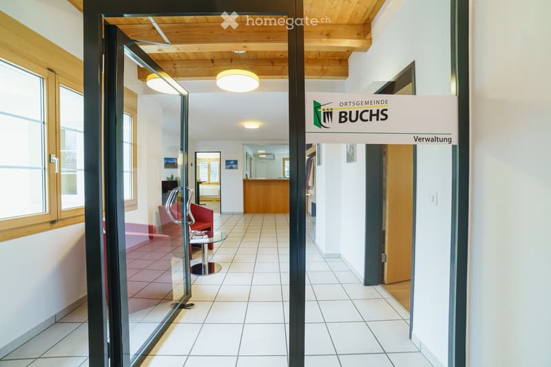 A modern hallway with a glass door leading to a reception area, featuring a red sofa, a glass table, and a chair.