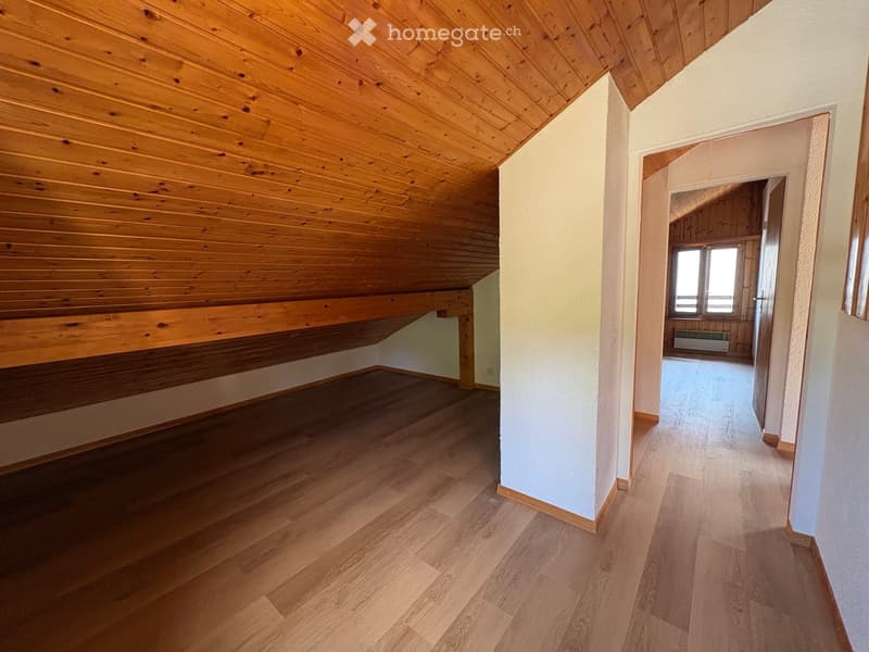 Empty room with wooden flooring and exposed wooden ceiling beams, featuring a doorway leading to another room with a window and radiator.