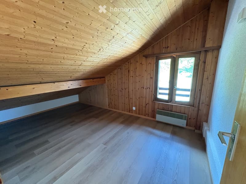 Empty attic room with wooden walls, ceiling, and floor, featuring a large window and a radiator.