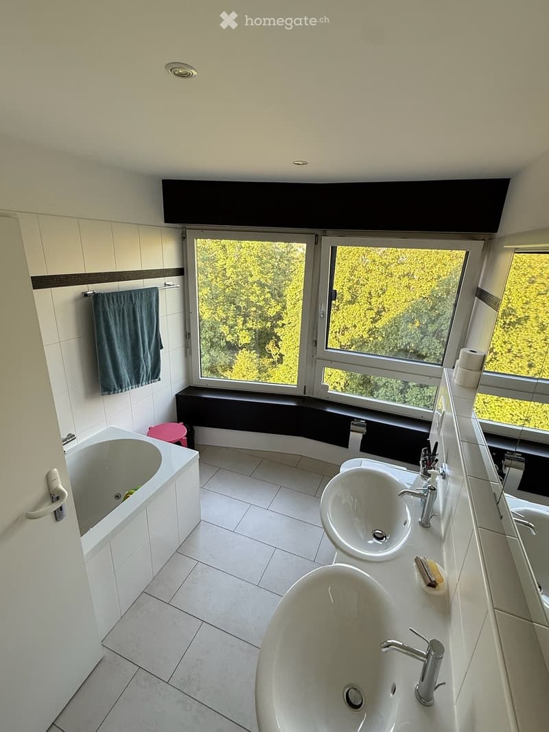 Modern bathroom with white tiles, two sinks, a bathtub, large windows, and a towel on the wall.