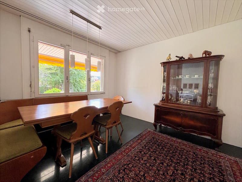 Bright dining area with a wooden table, chairs, a cabinet with glass doors, and a patterned rug on the floor.