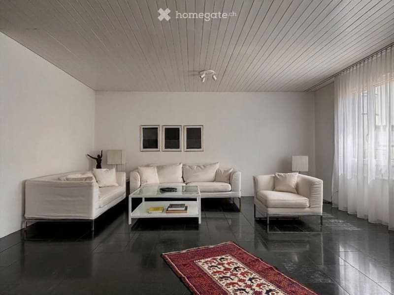 Modern living room with white walls, dark tiled floor, and large windows. Features a white sectional sofa, glass coffee table, armchairs, table lamps, and decorative picture frames.