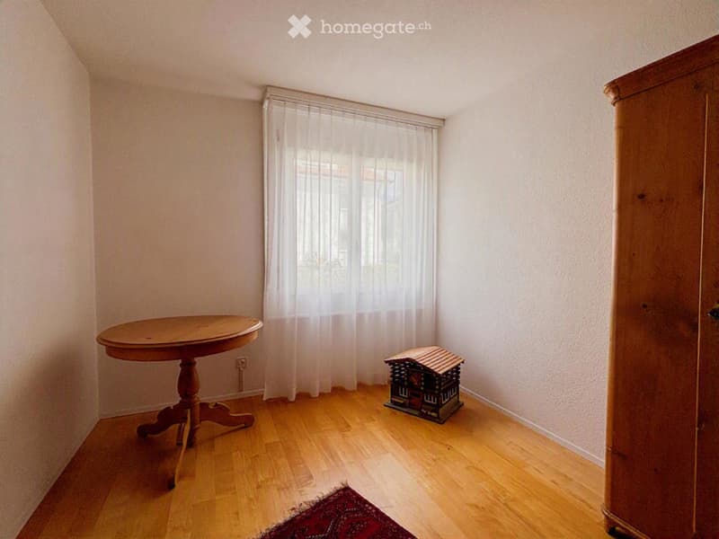 A small, unfurnished room with wooden flooring, a round wooden table, a large wardrobe, a small toy house, and a red rug near the window.