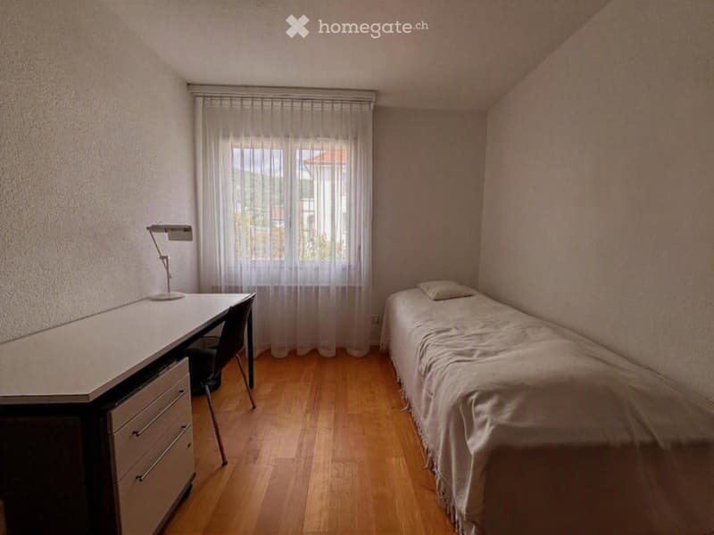 Cozy bedroom with wooden flooring, a single bed with white bedding, a desk with a chair, and a lamp on the desk.