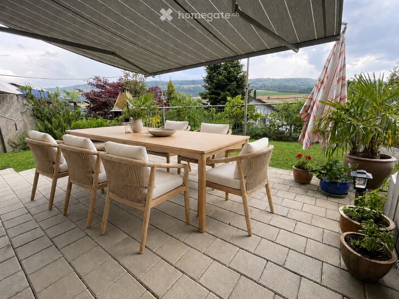 A modern outdoor patio with a wooden table, six wicker chairs, a large umbrella, and several potted plants, overlooking a scenic landscape.
