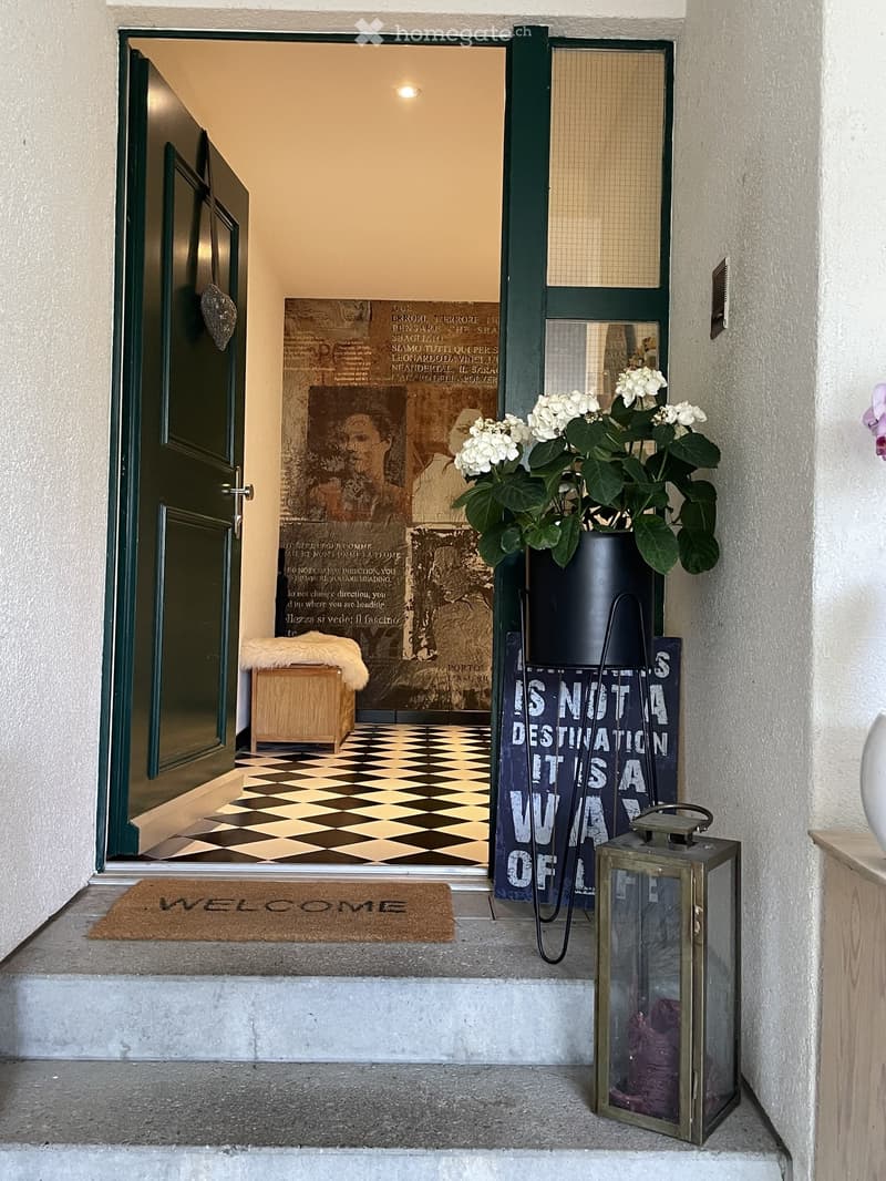 Entrance area with a checkered floor, a welcome mat, a decorative sign, a lantern, and a vase with white flowers.