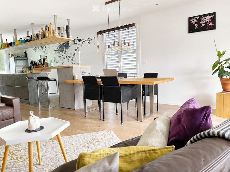 Modern open-plan living and dining area with a sofa, coffee table, dining table with chairs, bar stools, and a kitchen area with shelves and appliances.
