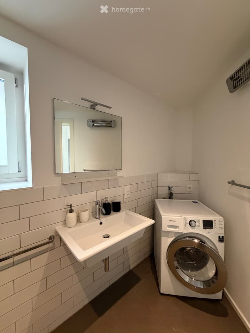 Bathroom. Modern bathroom with a white sink, mirror, and a Samsung washing machine. The walls are tiled in white, and there is a window providing natural light.
