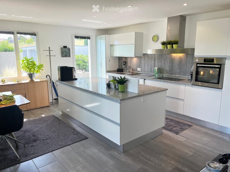 Modern kitchen with white cabinets, granite countertops, stainless steel appliances, and a central island. Features include a sink, coffee maker, oven, and potted plants on the windowsill and island.